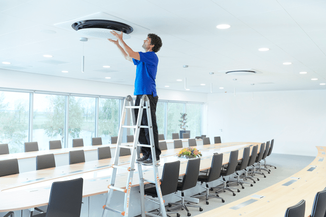 Samsung trained installer in blue polo shirt installing an 360 cassette air conditioner on a ladder in a conference room